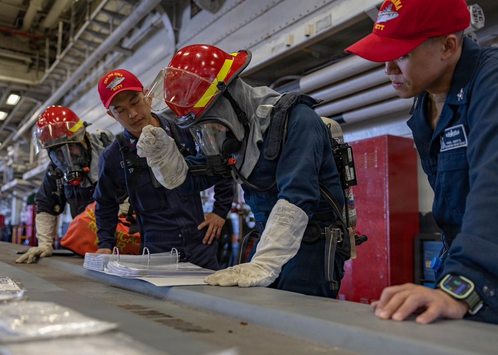 Military personnel aboard what appears to be a naval vessel are engaged in a damage control or firefighting drill. Several crew members wearing red helmets labeled 'IWO JIMA DCTT' (Damage Control Training Team) are reviewing documents in a binder on a table. One crew member is equipped with full firefighting gear including a breathing apparatus (SCBA), protective helmet with face shield, and heat-resistant gloves.