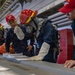 Military personnel aboard what appears to be a naval vessel are engaged in a damage control or firefighting drill. Several crew members wearing red helmets labeled 'IWO JIMA DCTT' (Damage Control Training Team) are reviewing documents in a binder on a table. One crew member is equipped with full firefighting gear including a breathing apparatus (SCBA), protective helmet with face shield, and heat-resistant gloves.