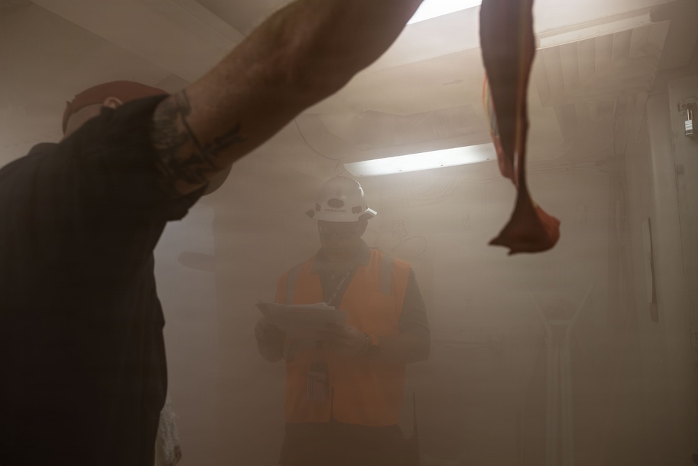 Two workers are visible in a dust-filled interior space, likely a construction or demolition site. One worker in the foreground wears a red hard hat and is raising their arm, while a second worker in the background wears a white hard hat, orange high-visibility vest, and face covering while holding a clipboard or tablet. The environment is heavily obscured by airborne dust or particulate matter.