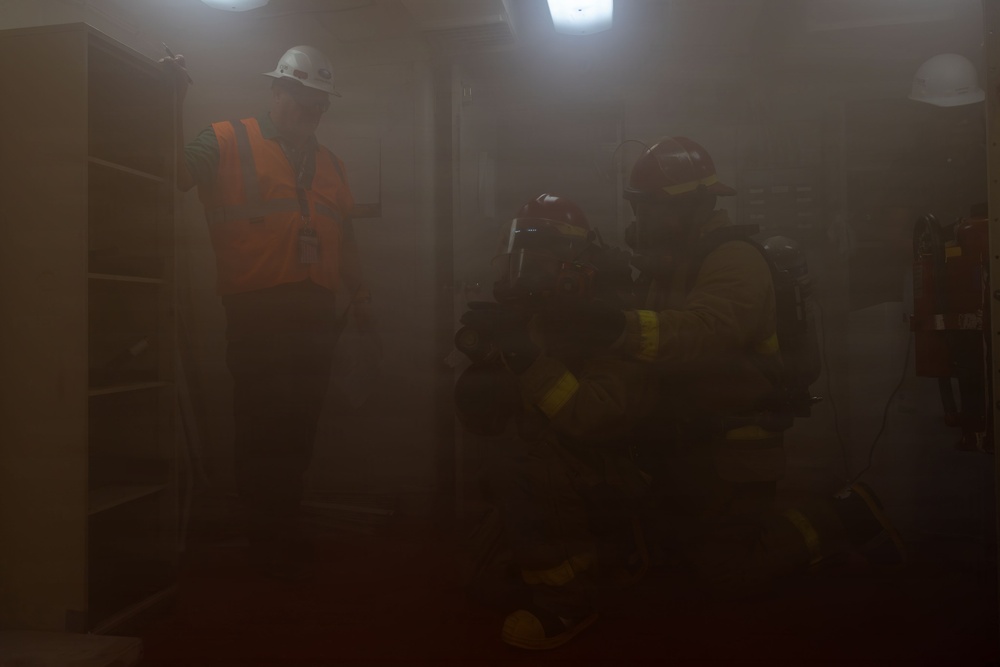 The image depicts what appears to be a fire training or emergency response exercise taking place in a smoke-filled indoor environment. A person in an orange high-visibility vest and white hard hat stands to the left, while two individuals in full firefighter turnout gear and helmets crouch low on the right, consistent with low-visibility search procedures. The heavy smoke significantly reduces visibility throughout the scene.