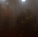 The image depicts what appears to be a fire training or emergency response exercise taking place in a smoke-filled indoor environment. A person in an orange high-visibility vest and white hard hat stands to the left, while two individuals in full firefighter turnout gear and helmets crouch low on the right, consistent with low-visibility search procedures. The heavy smoke significantly reduces visibility throughout the scene.