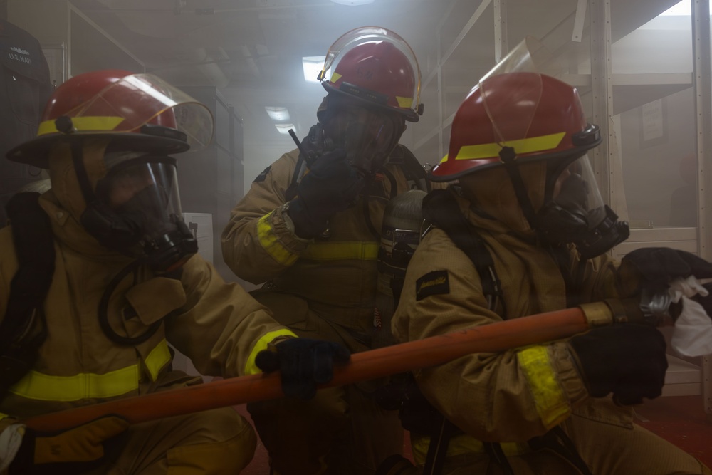 Three firefighters in full protective gear are operating inside a smoke-filled enclosed space, consistent with a U.S. Navy shipboard firefighting drill or actual emergency response. They are wearing tan/gold proximity suits, red helmets with yellow reflective bands, and full-face respirator masks. At least one firefighter is holding an orange fire hose, and another appears to be communicating via radio.