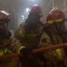 Three firefighters in full protective gear are operating inside a smoke-filled enclosed space, consistent with a U.S. Navy shipboard firefighting drill or actual emergency response. They are wearing tan/gold proximity suits, red helmets with yellow reflective bands, and full-face respirator masks. At least one firefighter is holding an orange fire hose, and another appears to be communicating via radio.