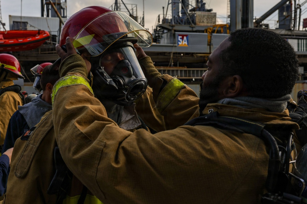 Two individuals in firefighting gear are shown on what appears to be a naval vessel or industrial dock, with one person assisting the other in securing a red helmet with a face shield and breathing apparatus. The scene suggests a firefighting drill or equipment check in a maritime environment. Additional personnel in similar gear are visible in the background.