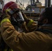 Two individuals in firefighting gear are shown on what appears to be a naval vessel or industrial dock, with one person assisting the other in securing a red helmet with a face shield and breathing apparatus. The scene suggests a firefighting drill or equipment check in a maritime environment. Additional personnel in similar gear are visible in the background.