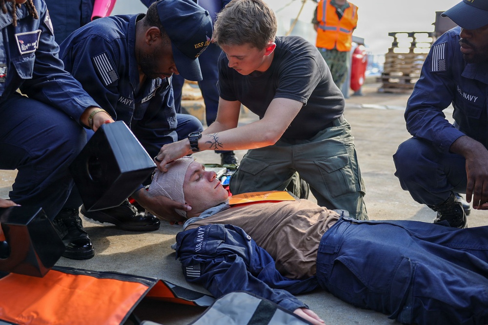 U.S. Navy personnel are conducting a medical training exercise on what appears to be a ship's deck or pier. A simulated casualty is lying on the ground with head bandaging applied and a cervical collar in place, while two sailors attend to the patient. The scene includes medical equipment and additional personnel observing, consistent with a damage control or first aid drill.