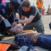 U.S. Navy personnel are conducting a medical training exercise on what appears to be a ship's deck or pier. A simulated casualty is lying on the ground with head bandaging applied and a cervical collar in place, while two sailors attend to the patient. The scene includes medical equipment and additional personnel observing, consistent with a damage control or first aid drill.
