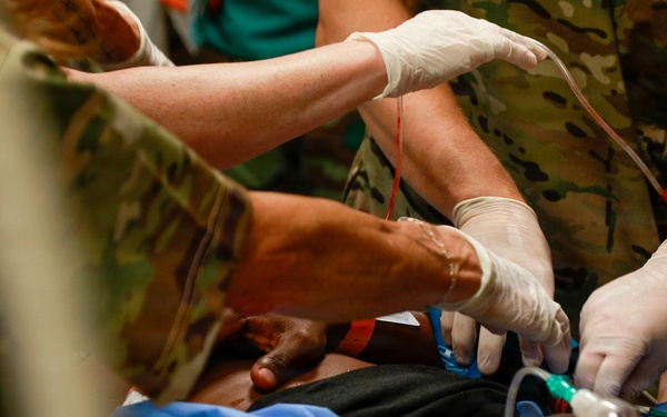 Military medical personnel in camouflage uniforms are providing emergency medical care to a civilian patient. Multiple gloved hands are visible administering treatment, including what appears to be IV line placement and oxygen mask application. The scene depicts an urgent tactical or field medical response.