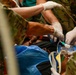 Military medical personnel in camouflage uniforms are providing emergency medical care to a civilian patient. Multiple gloved hands are visible administering treatment, including what appears to be IV line placement and oxygen mask application. The scene depicts an urgent tactical or field medical response.
