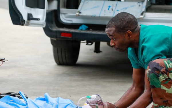 Two medical personnel wearing green scrubs and gloves are providing emergency care to an injured patient lying on the ground. The patient appears to have a bandaged lower limb wound and is receiving oxygen via a face mask. The scene takes place outdoors near a vehicle, with visible blood on the ground indicating a serious trauma situation.