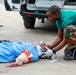 Two medical personnel wearing green scrubs and gloves are providing emergency care to an injured patient lying on the ground. The patient appears to have a bandaged lower limb wound and is receiving oxygen via a face mask. The scene takes place outdoors near a vehicle, with visible blood on the ground indicating a serious trauma situation.
