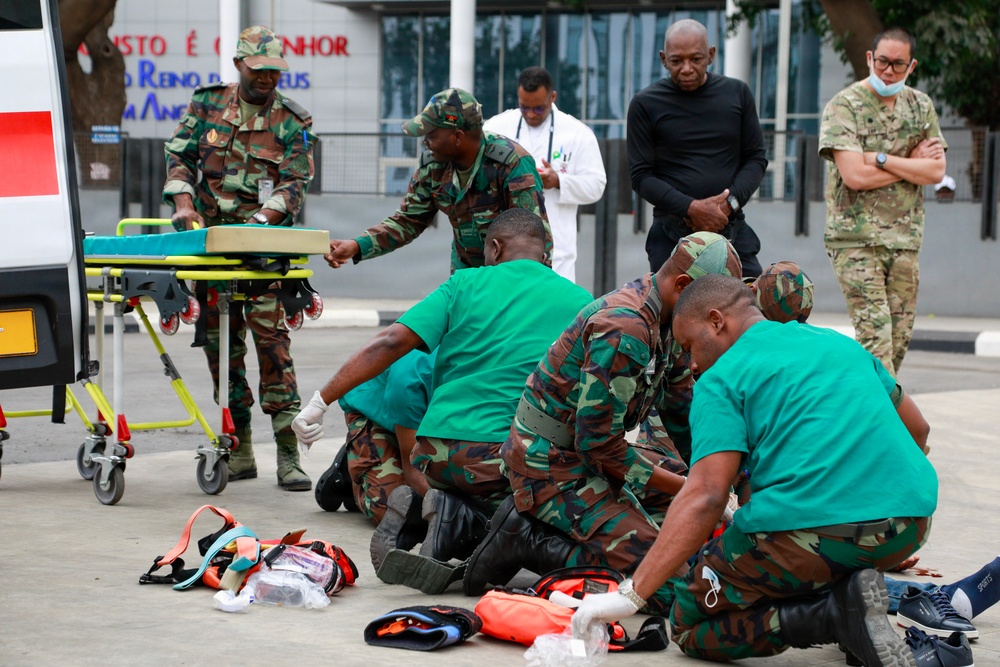 A group of military medical personnel in camouflage uniforms and green scrubs are conducting what appears to be a medical training exercise or emergency response drill outdoors. Several individuals are attending to a simulated casualty on the ground while observers, including a person in a white lab coat and another in US-pattern multicam uniform, watch the procedure. Medical equipment including a stretcher, orange medical bags, and other supplies are visible on the ground.