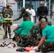 A group of military medical personnel in camouflage uniforms and green scrubs are conducting what appears to be a medical training exercise or emergency response drill outdoors. Several individuals are attending to a simulated casualty on the ground while observers, including a person in a white lab coat and another in US-pattern multicam uniform, watch the procedure. Medical equipment including a stretcher, orange medical bags, and other supplies are visible on the ground.