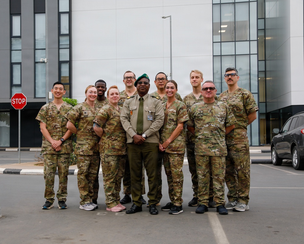 A group of approximately ten military personnel pose for a formal group photograph in a parking lot in front of a modern multi-story building. The majority are wearing US Army MultiCam (OCP) camouflage uniforms, while one individual in the center wears a distinct olive/khaki dress uniform with a green beret, suggesting a different national military affiliation. The setting appears to be an official or institutional facility, likely during a joint military engagement or training event.