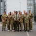 A group of approximately ten military personnel pose for a formal group photograph in a parking lot in front of a modern multi-story building. The majority are wearing US Army MultiCam (OCP) camouflage uniforms, while one individual in the center wears a distinct olive/khaki dress uniform with a green beret, suggesting a different national military affiliation. The setting appears to be an official or institutional facility, likely during a joint military engagement or training event.