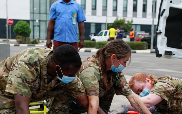 Military medical personnel in camouflage uniforms and surgical masks are conducting what appears to be a mass casualty training exercise outdoors. Four uniformed responders are attending to a simulated casualty on a blue stretcher mat, with visible simulated wound makeup on the patient's legs and head. A yellow stretcher, ambulance, and civilian observers are visible in the background.