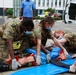 Military medical personnel in camouflage uniforms and surgical masks are conducting what appears to be a mass casualty training exercise outdoors. Four uniformed responders are attending to a simulated casualty on a blue stretcher mat, with visible simulated wound makeup on the patient's legs and head. A yellow stretcher, ambulance, and civilian observers are visible in the background.