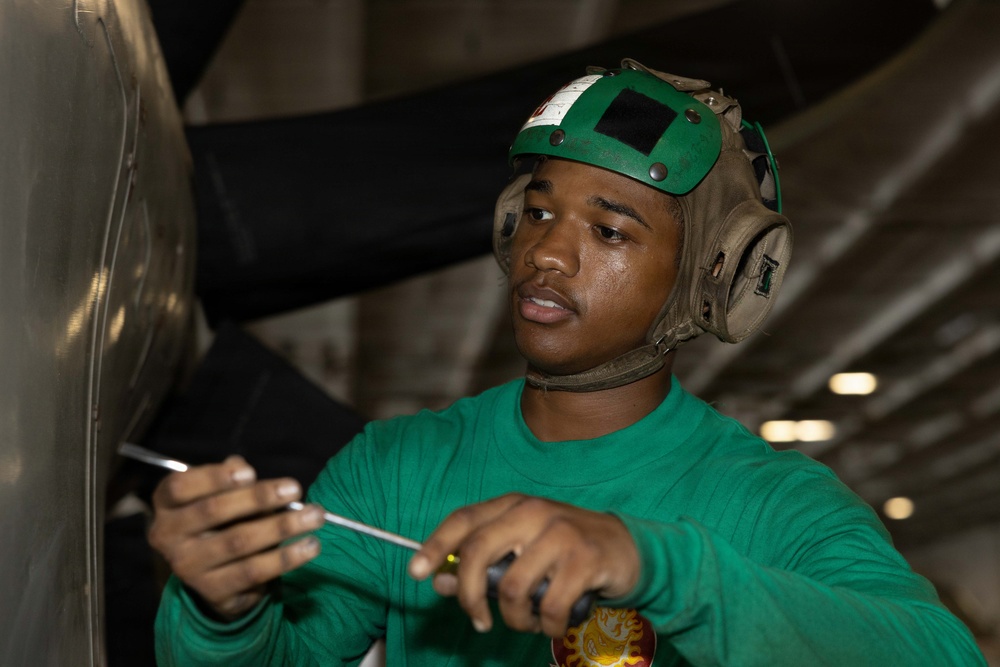 A young male technician wearing a green jersey and a green flight deck helmet is performing maintenance work using screwdrivers on what appears to be aircraft equipment. The green jersey and helmet are consistent with U.S. Navy aircraft carrier flight deck crew, where green designates aviation maintenance personnel. The setting appears to be inside a hangar bay or below-deck maintenance area.