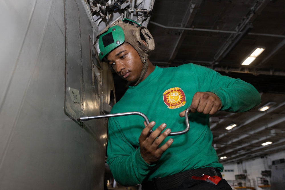 A military personnel member wearing a green long-sleeve shirt and a green flight deck helmet is using an L-shaped wrench or tool on what appears to be aircraft or ship equipment. The setting appears to be inside a naval vessel's hangar bay, indicated by the industrial ceiling, lighting, and metal structures visible in the background. The individual is focused on performing a maintenance or mechanical task.