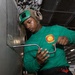 A military personnel member wearing a green long-sleeve shirt and a green flight deck helmet is using an L-shaped wrench or tool on what appears to be aircraft or ship equipment. The setting appears to be inside a naval vessel's hangar bay, indicated by the industrial ceiling, lighting, and metal structures visible in the background. The individual is focused on performing a maintenance or mechanical task.
