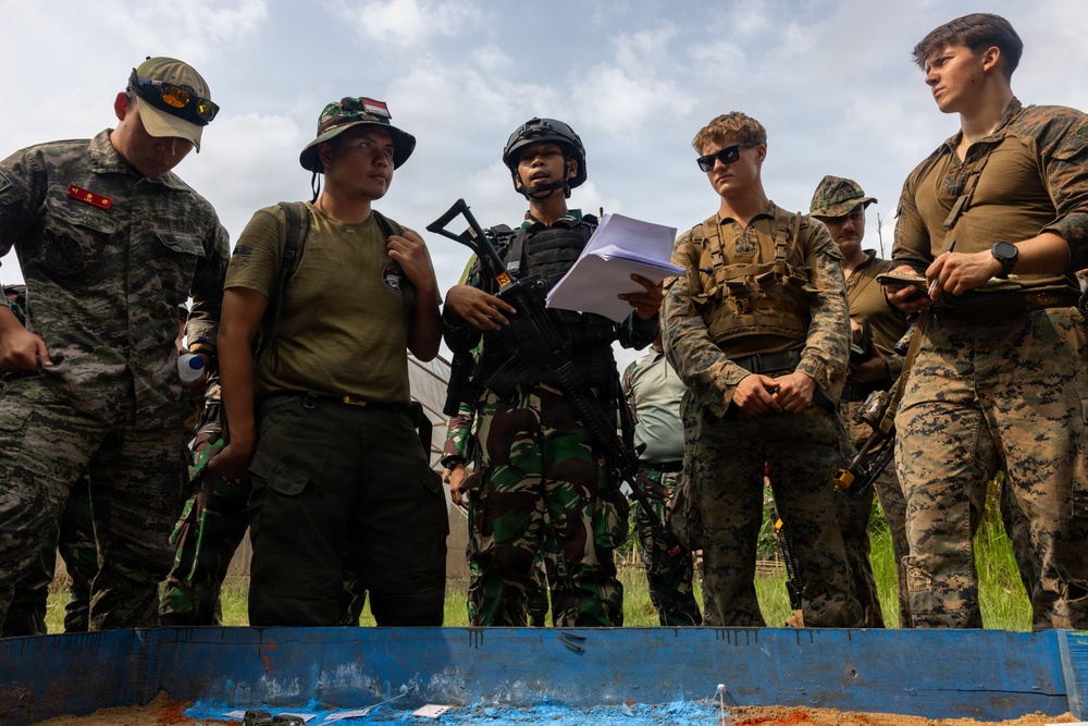A group of military personnel from multiple nations are gathered around a blue table or surface, appearing to review materials during what looks like a joint military exercise or briefing. The group includes soldiers in different camouflage patterns, suggesting a multinational training event. One soldier in the center holds papers and carries a rifle while wearing a tactical helmet and vest.