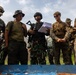 A group of military personnel from multiple nations are gathered around a blue table or surface, appearing to review materials during what looks like a joint military exercise or briefing. The group includes soldiers in different camouflage patterns, suggesting a multinational training event. One soldier in the center holds papers and carries a rifle while wearing a tactical helmet and vest.