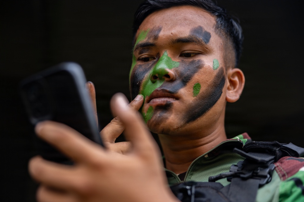 A young male soldier wearing military camouflage uniform and tactical vest is applying green and black face paint while using a smartphone as a mirror. The subject has Southeast Asian features and is focused on his reflection in the phone screen. The image is a close-up portrait with a dark, blurred background.