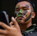 A young male soldier wearing military camouflage uniform and tactical vest is applying green and black face paint while using a smartphone as a mirror. The subject has Southeast Asian features and is focused on his reflection in the phone screen. The image is a close-up portrait with a dark, blurred background.
