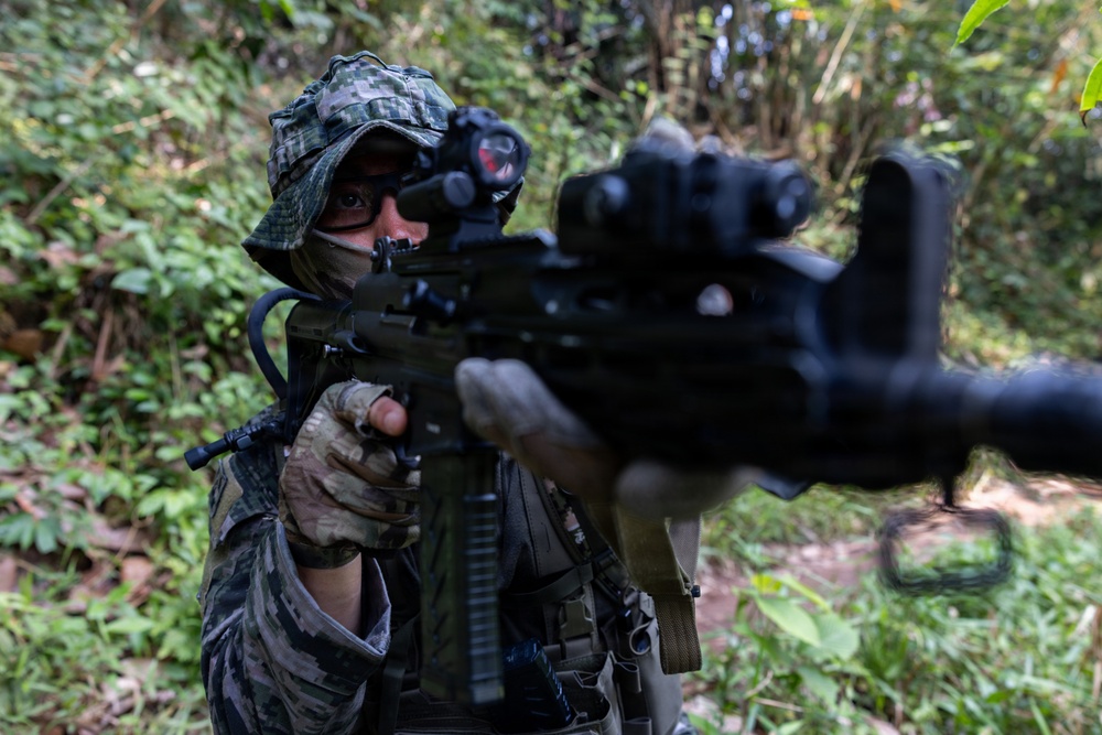 A soldier or tactical operator in full camouflage gear is aiming a rifle directly toward the camera in a dense jungle or forest environment. The individual is wearing a camouflage hood, face covering, tactical gloves, and has an optic sight mounted on the weapon. The close-up perspective creates a dramatic, immersive composition with the muzzle of the weapon in the foreground.