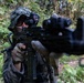 A soldier or tactical operator in full camouflage gear is aiming a rifle directly toward the camera in a dense jungle or forest environment. The individual is wearing a camouflage hood, face covering, tactical gloves, and has an optic sight mounted on the weapon. The close-up perspective creates a dramatic, immersive composition with the muzzle of the weapon in the foreground.