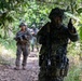The image shows a group of military personnel conducting a patrol or training exercise along a jungle trail. Multiple soldiers from what appear to be different units or nationalities are visible, with the foreground soldier in full tactical gear giving a hand signal. The setting is a dense tropical environment with fruit-bearing trees overhead.