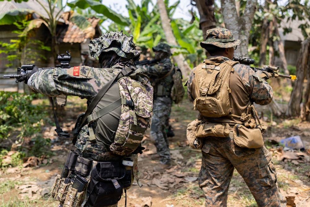 The image shows multiple armed military personnel in camouflage uniforms conducting what appears to be a tactical training exercise in a tropical or jungle environment. Soldiers are seen in combat-ready stances with weapons raised, suggesting a patrol or clearing operation drill. The setting includes vegetation, banana trees, and a rustic structure in the background.