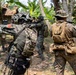 The image shows multiple armed military personnel in camouflage uniforms conducting what appears to be a tactical training exercise in a tropical or jungle environment. Soldiers are seen in combat-ready stances with weapons raised, suggesting a patrol or clearing operation drill. The setting includes vegetation, banana trees, and a rustic structure in the background.