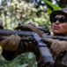 A soldier in military camouflage uniform is positioned in a jungle or dense forest environment, holding a rifle in a ready position. The individual is wearing tactical gear including a camouflage patrol cap, dark sunglasses, and tactical gloves. The image appears to depict a military patrol or training exercise in a tropical setting.