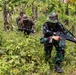 Three armed soldiers are conducting a patrol or training exercise through dense tropical jungle vegetation. Two soldiers in the background wear tan/brown camouflage uniforms consistent with US Marine Corps pattern, while the lead soldier wears a green woodland camouflage uniform and bucket hat consistent with Indonesian TNI (Tentara Nasional Indonesia) military dress. The setting and mixed uniforms suggest a joint military training exercise in a tropical environment.