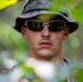 A young male soldier or military personnel is photographed in a forested environment, wearing camouflage gear. He is partially obscured by out-of-focus foliage in the foreground, suggesting a surveillance or patrol context. The close-up portrait emphasizes a serious, alert expression.