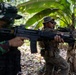 Two armed military personnel are conducting what appears to be a joint training exercise in a tropical jungle environment. One soldier wears Indonesian Army (TNI-AD) woodland camouflage uniform, while the other wears US Marine Corps MARPAT digital camouflage with tactical gear. Both are in a low ready or aimed firing stance among dense tropical vegetation including banana trees.