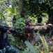The image shows multiple armed military personnel conducting what appears to be a joint training exercise in a dense jungle or tropical forest environment. At least two distinct camouflage patterns are visible, suggesting soldiers from different military units or nations are participating together. The personnel are in tactical positions with weapons raised, consistent with a patrol or combat training scenario.