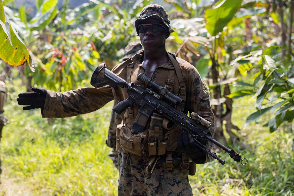 A soldier in digital camouflage uniform is pictured in a tropical jungle environment, carrying a scoped rifle and wearing full tactical gear. The soldier appears to be directing or signaling with an outstretched arm while on patrol. Additional personnel are visible in the background among dense tropical vegetation.