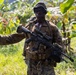 A soldier in digital camouflage uniform is pictured in a tropical jungle environment, carrying a scoped rifle and wearing full tactical gear. The soldier appears to be directing or signaling with an outstretched arm while on patrol. Additional personnel are visible in the background among dense tropical vegetation.