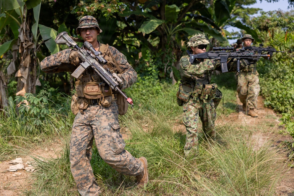 Three armed military personnel are conducting a patrol or training exercise in a tropical jungle environment. The lead soldier wears US Marine Corps MARPAT digital camouflage and carries a scoped rifle, while two other soldiers in different camouflage patterns follow behind. The setting and mixed uniforms suggest a multinational joint military exercise or training operation.