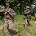 Three armed military personnel are conducting a patrol or training exercise in a tropical jungle environment. The lead soldier wears US Marine Corps MARPAT digital camouflage and carries a scoped rifle, while two other soldiers in different camouflage patterns follow behind. The setting and mixed uniforms suggest a multinational joint military exercise or training operation.