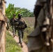 A group of military personnel in woodland camouflage uniforms are conducting a patrol along a narrow dirt path between what appear to be dried palm frond structures. The lead soldier is carrying an assault rifle at the ready while moving through a rural, tropical environment. The setting suggests a training exercise or operational patrol in a tropical region, evidenced by banana trees and agricultural fields in the background.