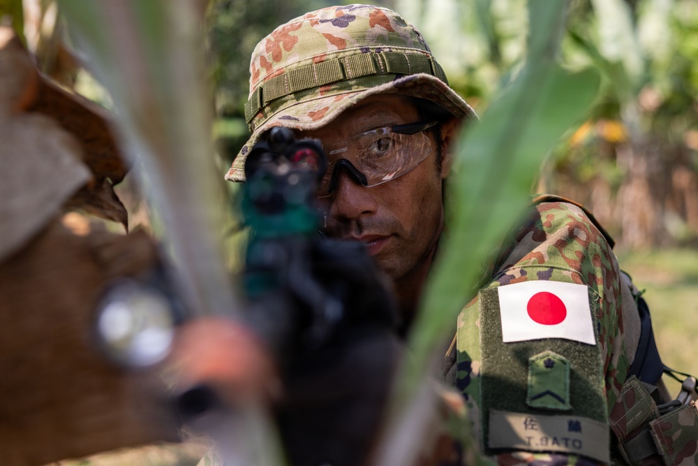 A Japan Ground Self-Defense Force (JGSDF) soldier is photographed in a tactical position among vegetation, aiming a weapon toward the camera. The soldier wears Japanese camouflage uniform with a Japanese national flag patch and a name tape reading 'T. SATO' (佐藤). The image appears to be taken during a military exercise or training operation.