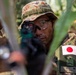 A Japan Ground Self-Defense Force (JGSDF) soldier is photographed in a tactical position among vegetation, aiming a weapon toward the camera. The soldier wears Japanese camouflage uniform with a Japanese national flag patch and a name tape reading 'T. SATO' (佐藤). The image appears to be taken during a military exercise or training operation.