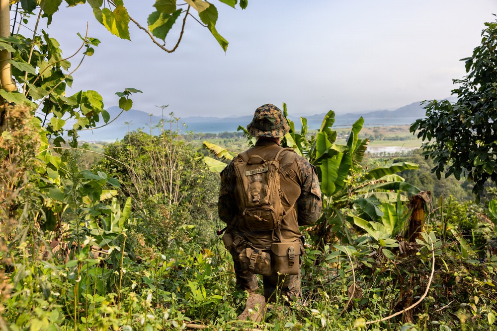 A U.S. Marine in full camouflage gear stands on an elevated jungle hillside, looking out over a tropical valley with a body of water and mountains in the background. The soldier is viewed from behind, wearing MARPAT woodland camouflage, a boonie hat, and carrying a tactical backpack with a visible name tape reading 'GODDARD'. The lush tropical vegetation, including banana trees, suggests a Southeast Asian or Pacific Island environment.