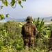 A U.S. Marine in full camouflage gear stands on an elevated jungle hillside, looking out over a tropical valley with a body of water and mountains in the background. The soldier is viewed from behind, wearing MARPAT woodland camouflage, a boonie hat, and carrying a tactical backpack with a visible name tape reading 'GODDARD'. The lush tropical vegetation, including banana trees, suggests a Southeast Asian or Pacific Island environment.