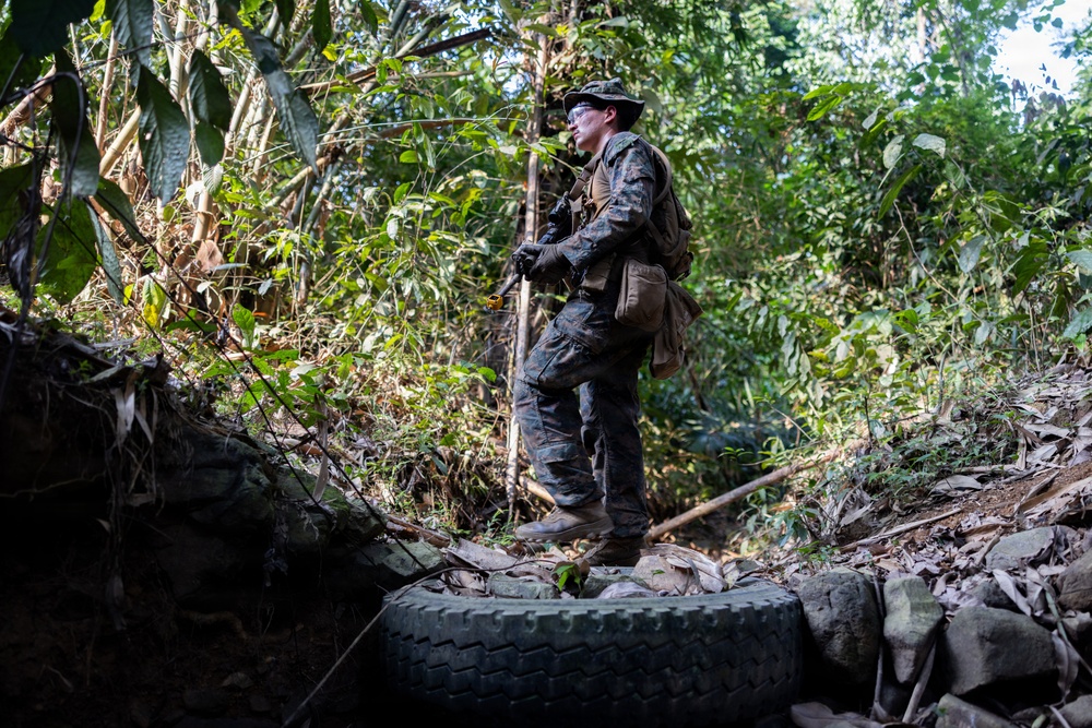 A soldier or military personnel in full camouflage gear stands on a discarded tire in a dense jungle or forest environment. The individual is equipped with tactical gear, gloves, protective eyewear, and a boonie hat, and appears to be scanning or observing the surrounding terrain. The setting suggests a field training exercise or patrol operation in a tropical environment.