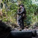A soldier or military personnel in full camouflage gear stands on a discarded tire in a dense jungle or forest environment. The individual is equipped with tactical gear, gloves, protective eyewear, and a boonie hat, and appears to be scanning or observing the surrounding terrain. The setting suggests a field training exercise or patrol operation in a tropical environment.