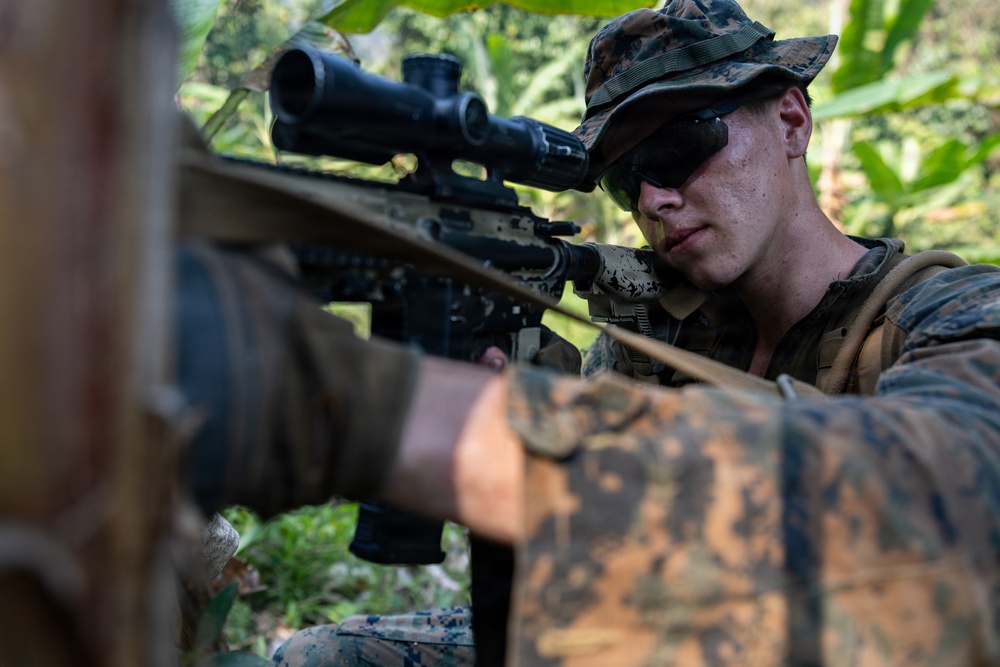 A young male soldier in MARPAT (Marine pattern) camouflage uniform is aiming a scoped rifle in a jungle or forested environment. He is wearing tactical eyewear pushed up on his face and a camouflage patrol cap. The image is taken from a low, close-up angle emphasizing the weapon and the shooter's focused expression.